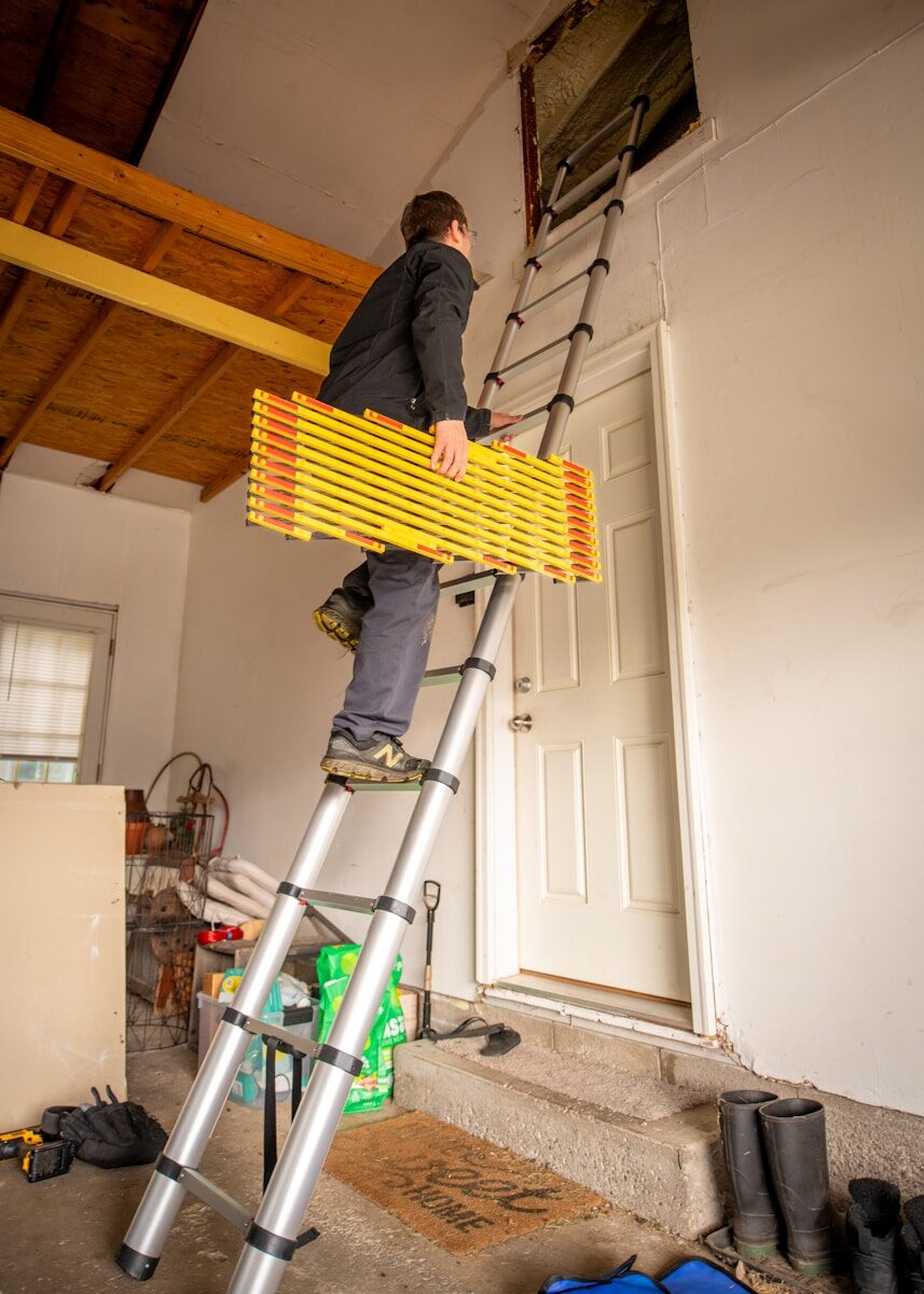 A person climbs a ladder while carrying a folding device towards an overhead entrance in a garage, illustrating home maintenance or repair activities.